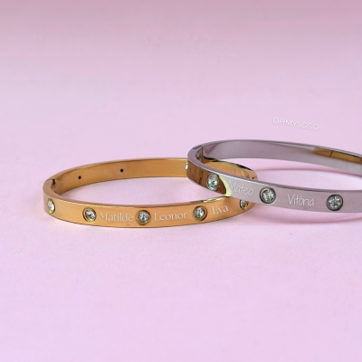 Dois braceletes metálicos, dourado e prateado, com pedras e nomes gravados em fundo cor-de-rosa.