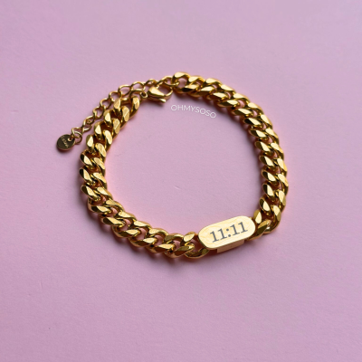 Pulseira dourada com placa numerada 11:11 sobre fundo rosa
