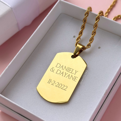 Colar dourado com pendente gravado em caixa branca sobre fundo rosa