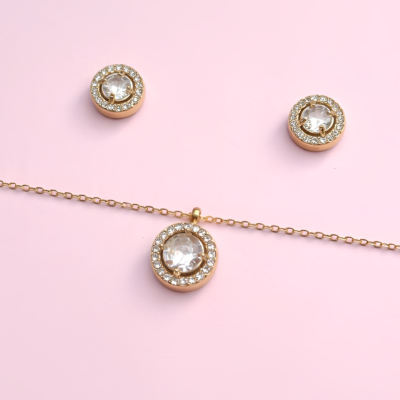 Conjunto de joalharia dourado com cristais transparentes em fundo rosa.