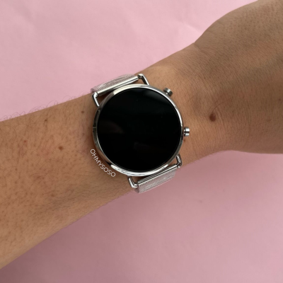 Relógio inteligente prateado com pulseira metálica num pulso sobre fundo rosa