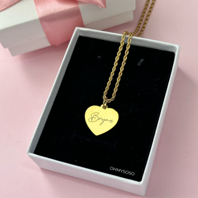 Colar dourado com pendente coração gravado com o nome Bryan numa caixa branca com interior preto e caixa com laço cor de rosa ao fundo