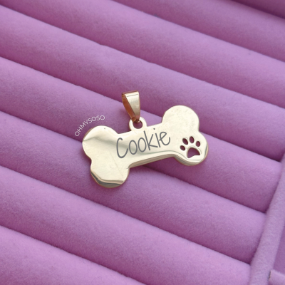 Pendente dourado em forma de osso com nome 'Cookie' em fundo lilás almofadado