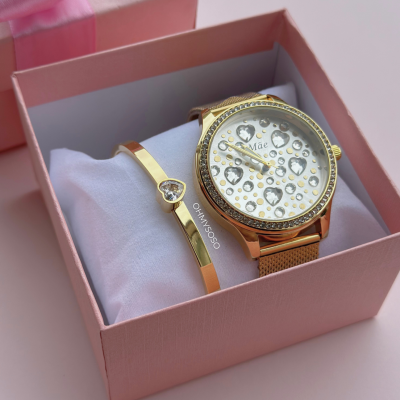 Relógio dourado com mostrador branco e corações com texto 'Mãe' e bracelete dourada com coração cravejado em caixa rosa.