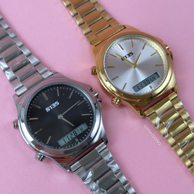 Dois relógios de pulso metálicos, um prata e outro dourado, com mostradores analógicos e visor digital, sobre fundo rosa.