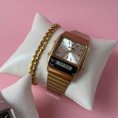 Relógio dourado digital e analógico com pulseira metálica e pulseira de elos dourados sobre almofada branca, fundo rosa