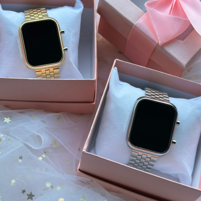 Relógios inteligentes dourado e prateado em caixas cor-de-rosa sobre tecido com estrelas douradas.