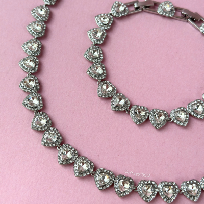 Colar e pulseira prateados com pedras em forma de coração transparentes sobre fundo rosa