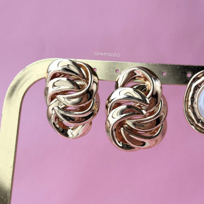 Brincos dourados entrelaçados pendurados numa estrutura metálica, fundo rosa