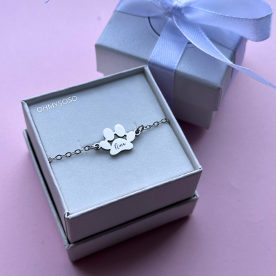 Pulseira prateada com pendente em forma de pata e nome 'Nina' gravado, em caixa branca com laço branco