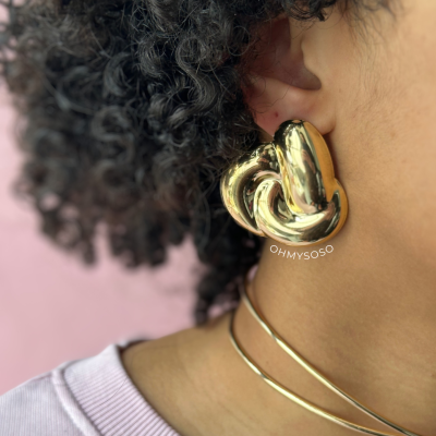 Brinco trançado dourado num ouvido com fundo rosa e colares dourados