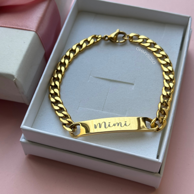 Pulseira dourada com placa gravada 'Mimi' dentro de caixa branca sobre fundo rosa.