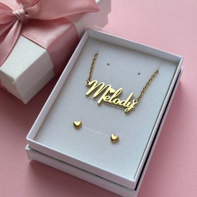 Conjunto de joalharia dourada com colar 'Melody' e brincos em forma de coração numa caixa branca com laço rosa