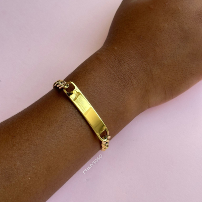 Pulseira dourada com placa lisa num pulso sobre fundo rosa claro