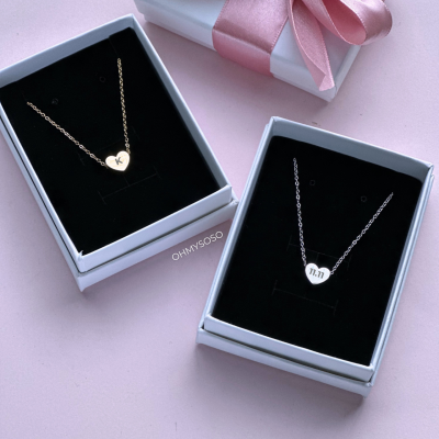 Dois colares com pendentes em forma de coração em caixas brancas, com letra 'K' dourada e números '11:11' prateados, com caixa branca e fita rosa ao lado