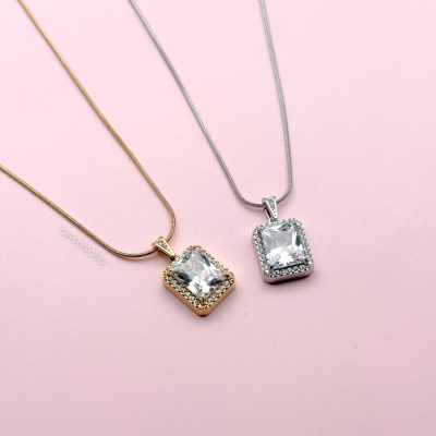 Duas colares com pendentes quadrados com pedras transparentes, um em dourado e outro em prateado, sobre fundo rosa.