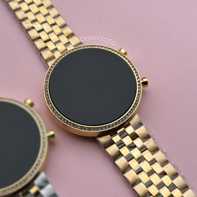 Dois relógios digitais com braceletes metálicos dourado e prateado, aro com pedras pretas, sobre fundo rosa.