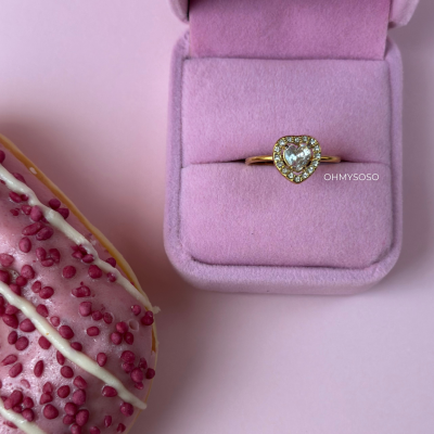 Anel dourado com pedra em forma de coração numa caixa rosa, ao lado de um donut glacê rosa