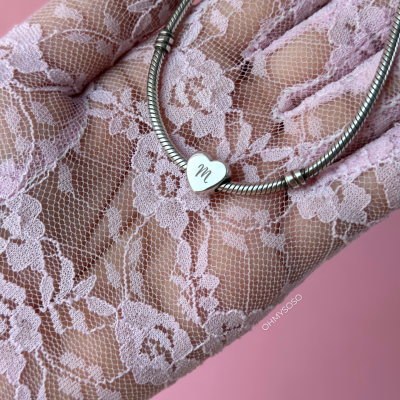 Pulseira prateada com coração gravado 'm' em mão com luva de renda rosa