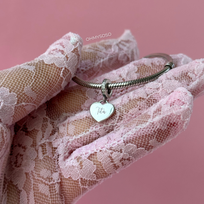 Pulseira prateada com pendente em formato de coração com o nome 'Tita' segurada por mão com luva de renda rosa