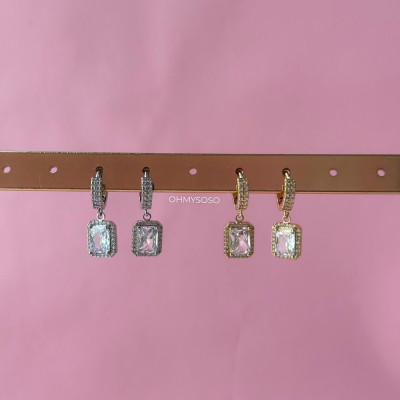 Dois pares de brincos prata e dourado com pedras quadradas pendurados numa fita castanha sobre fundo rosa