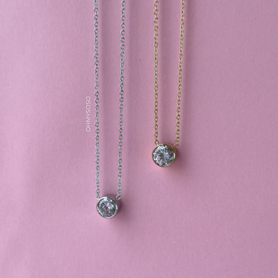 Dois colares finos com pendentes redondos em cristal, um prata e outro dourado, sobre fundo rosa.