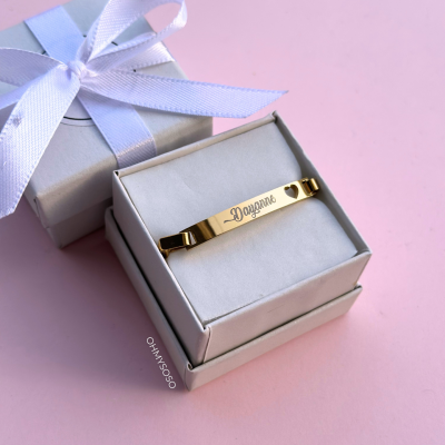 Pulseira dourada gravada numa caixa branca com laço prata sobre fundo rosa