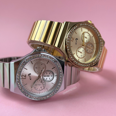 Dois relógios metálicos, um dourado e outro prateado, com cristais na moldura, em fundo rosa.