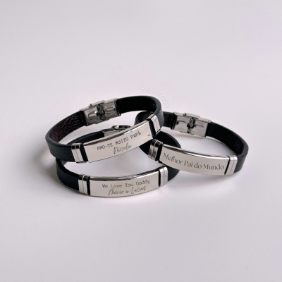 Três pulseiras pretas com placas metálicas gravadas, sobre fundo branco.