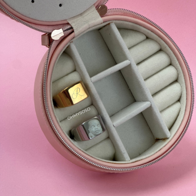 Estojo de joias redondo rosa com veludo cinza e dois anéis sinete, fundo rosa.