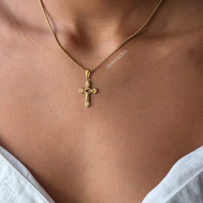 Colar dourado com pingente de cruz detalhada, usado por pessoa com vestido branco.
