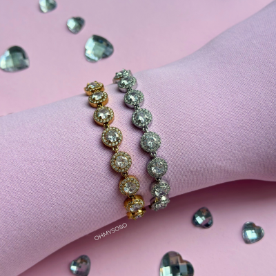 Dois braceletes com pedras brilhantes em dourado e prateado sobre tecido rosa claro com corações transparentes ao redor.