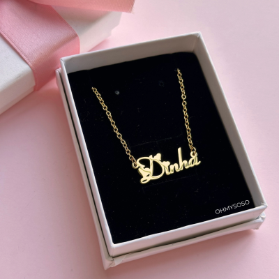 Colar dourado com escrita Dinha em caixa preta e branca com fita cor-de-rosa