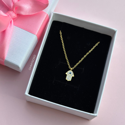 Colar dourado com pendente de mão Hamsá com pedras azuis e brancas numa caixa branca com fita rosa
