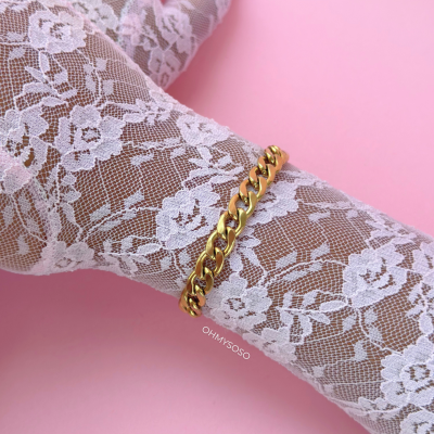 Pulseira dourada com elos grossos numa mão com luva de renda branca sobre fundo rosa.