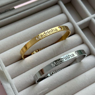 Duas pulseiras metálicas, dourada e prateada, gravadas com 'Madrinha' com um coração, numa caixa de veludo cinza.