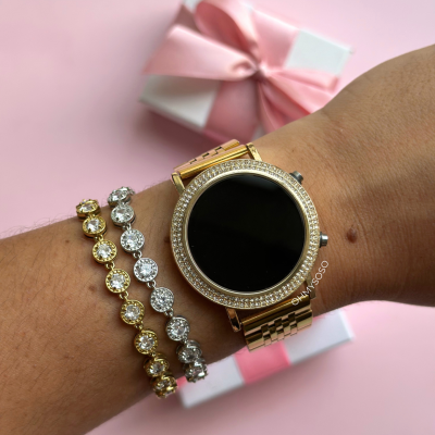 Relógio dourado com cristais e duas pulseiras com pedras em fundo rosa e caixa de presente