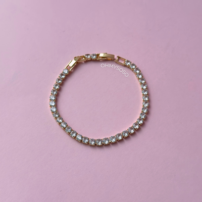 Pulseira tennis dourada com pedras brilhantes em fundo rosa.