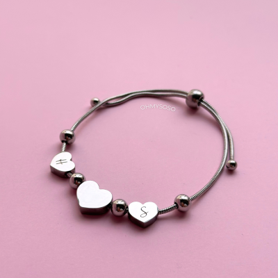 Pulseira metálica prateada com pendentes em forma de coração e letras, em fundo rosa