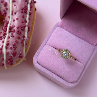 Anel de ouro numa caixa rosa ao lado de uma rosquinha com cobertura rosa