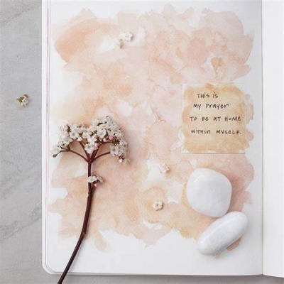 Caderno aberto com fundo rosa claro, flor seca branca, duas pedras brancas e papel com texto manuscrito em inglês.