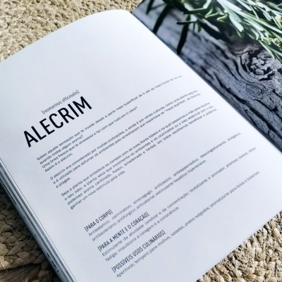 Livro aberto com texto sobre alecrim e foto da planta em superfície têxtil bege