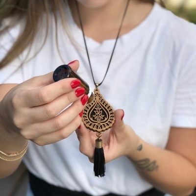 Colar de madeira com padrão gravado em forma de gota e borla preta, segurado por uma mulher com unhas vermelhas e t-shirt branca.