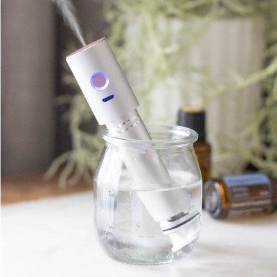 Difusor portátil branco em copo de vidro com água e vapor saindo, frascos ao fundo