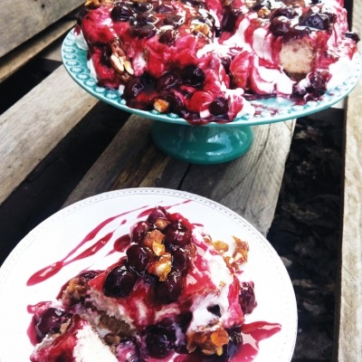 Bolo com cobertura de frutas vermelhas e nozes, servido num prato branco e suporte azul sobre madeira.