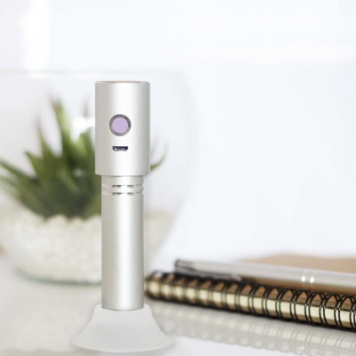 Dispositivo cilíndrico prateado com botão roxo sobre base branca, caderno espiral e planta ao fundo