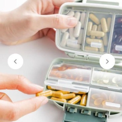 Caixinha verde com cápsulas brancas, bege e amarelas sendo aberta por duas mãos