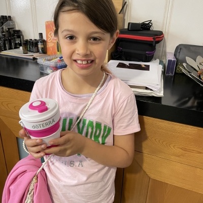 Menina de camiseta rosa segurando copo dōTERRA em cozinha com balcão e azulejos