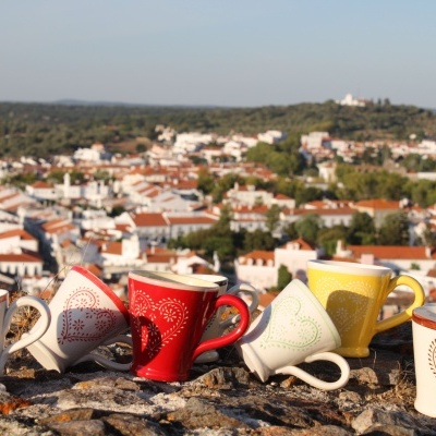 Conjunto de seis chávenas de cerâmica coloridas com padrão de corações, sobre pedra, com cidade ao fundo.