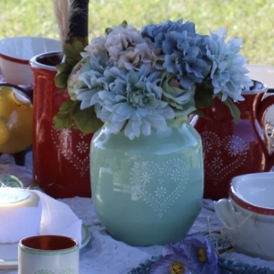 Vaso cerâmica verde com flores artificiais azuis e bege em mesa com louça variada.